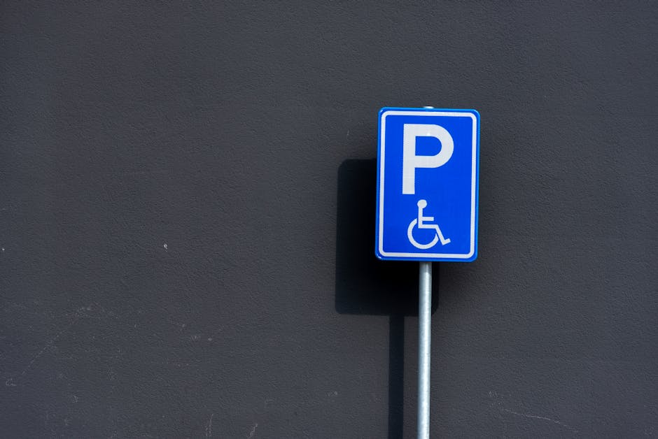 A close-up image of a blue parking sign with a white 'P' and a wheelchair symbol, mounted on a metal pole positioned against a dark grey textured wall. The sign indicates designated parking for disabled individuals, located on a paved area that is accessible for vehicles involved in home relocation or furniture transport. The shadow of the sign is cast on the wall, revealing daylight conditions. This setting may be near a property where removals or moving services, such as those offered by Movers Kentish Town, facilitate household moves within the Camden area, including parking permits for efficient loading and unloading of furniture and boxes, with equipment like trolleys and loading blankets potentially present nearby.