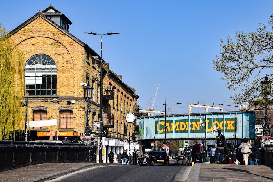 A busy scene outside Camden Town Underground Station with a diverse group of pedestrians on the pavement, some carrying backpacks and shopping bags, while others are waiting or walking towards the station entrance. The station's red brick façade features arched windows and a prominent blue sign reading 'Camden Town Station.' An underground roundel logo is mounted above the entrance. Nearby, a wooden bench is placed on the sidewalk. This setting is typical of urban house removal logistics, where furniture and moving boxes from local residences managed by Movers Kentish Town are likely being transported to or from the station area, supporting efficient home relocation and furniture transport services. The scene is illuminated by daylight, capturing the active movement of people involved in moving activities in Camden Town.