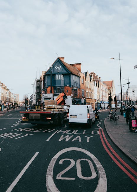 A flatbed truck parked on a street in Kentish Town, loaded with wooden pallets, cardboard boxes, and wrapped furniture pieces, prepared for a home relocation. The truck is positioned near a row of multi-storey brick buildings with shop fronts, while a white van is parked alongside it, possibly assisting in the moving process. The scene shows the loading area on a paved road with clear traffic markings, including a 20 mph speed limit sign painted on the road surface. Nearby, pedestrians and cyclists are visible on the sidewalk, with a lamppost and streetlights lining the street. The sky is partly cloudy, and the surroundings suggest a busy urban environment where furniture transport and packing are in progress, as part of professional removal services offered by Movers Kentish Town.