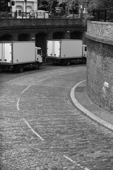 A black-and-white photograph captures a curve in a cobblestone driveway within an urban setting, featuring two white box trucks parked parallel to each other on the left side, with their rear doors facing an archway underneath a brick wall. The trucks are positioned close to a brick retaining wall on the right, which curves inward following the driveway’s bend. In the background, a railing and trees are visible above the structure, indicating a landscaped area or street level. The scene is illuminated by natural light, and the image suggests a home relocation or furniture transport process, with the trucks aligned for loading or unloading. The setting appears to be an area where Movers Kentish Town could assist with removals, packing, and moving services, especially given the focus on loading processes and vehicle positioning for efficient house removals.