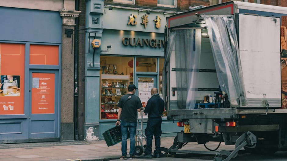 Two movers from Movers Kentish Town are preparing for a home relocation outside a Chinese restaurant named 'GUANG' in Kentish Town. One mover, holding a black crate, is standing beside a hand truck, while the other is near the open side of a large moving truck, which has a transparent plastic sheet partially covering its loading area. The truck is parked on the pavement just outside the restaurant, with the door closed. Inside the restaurant's large glass window, shelves with various items and signage are visible. The building's façade features ornate decorative moldings and signage in Chinese characters, with a mix of blue and beige colors. The scene is set during daytime with natural light illuminating the surroundings, capturing the process of packing and loading furniture and boxes as part of a furniture transport or house move. This image relates to the services provided by Movers Kentish Town, including removals and home relocation logistics, as discussed in the page about hidden fees before booking.