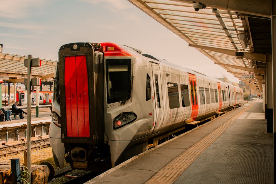 A modern silver and red train is stationary at a platform near Kentish Town West Station during daylight hours. The train features sleek, aerodynamic design with large windows and an identification number on the side. The platform has a textured yellow safety strip along the edge, with a metal roof canopy above protecting waiting passengers. In the background, there are a few seated passengers, some standing, and additional trains can be seen on adjacent tracks. Overhead lighting fixtures and station signage are visible under the canopy. The station environment appears clean and well-maintained, with natural light illuminating the scene, suggesting a typical day of home relocation planning involving train travel or transportation logistics, as provided by Movers Kentish Town specializing in removals and furniture transport services.