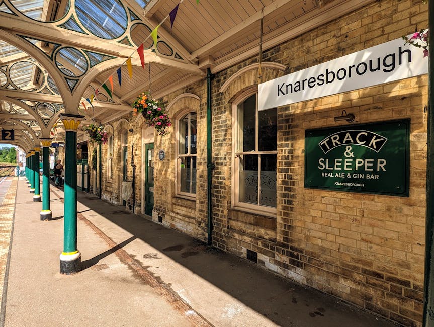 The image shows the platform of Knaresborough railway station with a row of green-painted metal columns supporting a decorative wooden canopy featuring intricate circular cutouts, under which hanging flower baskets are suspended. The platform is paved with concrete and runs alongside an old brick station building with arched windows framed in cream-colored trim. There are a few people in the distance, possibly waiting or preparing for a home relocation or moving service. Attached to the brick wall is a sign that reads 'Knaresborough' and a green and white sign for 'Track & Sleeper,' indicating a bar and pub located within the station premises. The scene is lit by natural sunlight, creating shadows on the platform, and the environment suggests an active setting for freight or passenger transport, suitable for a company like Movers Kentish Town that offers removals and furniture transport services near Kentish Town West Station.