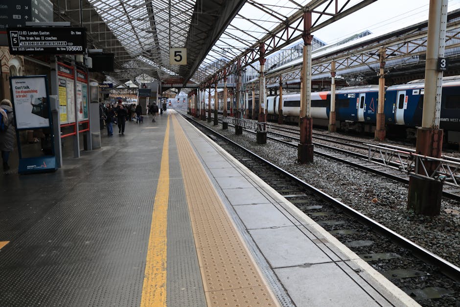 A modern silver and red train is stationary at a platform near Kentish Town West Station during daylight hours. The train features sleek, aerodynamic design with large windows and an identification number on the side. The platform has a textured yellow safety strip along the edge, with a metal roof canopy above protecting waiting passengers. In the background, there are a few seated passengers, some standing, and additional trains can be seen on adjacent tracks. Overhead lighting fixtures and station signage are visible under the canopy. The station environment appears clean and well-maintained, with natural light illuminating the scene, suggesting a typical day of home relocation planning involving train travel or transportation logistics, as provided by Movers Kentish Town specializing in removals and furniture transport services.