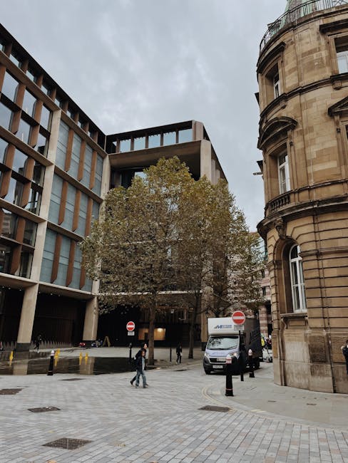 A street scene showing a modern multi-storey office building with large glass windows on the left and a historic curved sandstone building on the right near Kentish Town Road. In the center, there is a large tree with sparse green and brown leaves, indicating autumn. The street is paved with grey cobblestones, and several pedestrians are walking along the pavement. A white delivery van and a black car are parked near the buildings, with the van belonging to Movers Kentish Town, a professional removals company. Behind the vehicles, a loading area is visible with some caution cones and a few people standing nearby, possibly preparing for a house or office relocation. The overcast sky provides diffuse lighting, contributing to a neutral, daytime setting focused on urban moving logistics and furniture transport preparation in the vicinity of businesses and offices on Kentish Town Road.