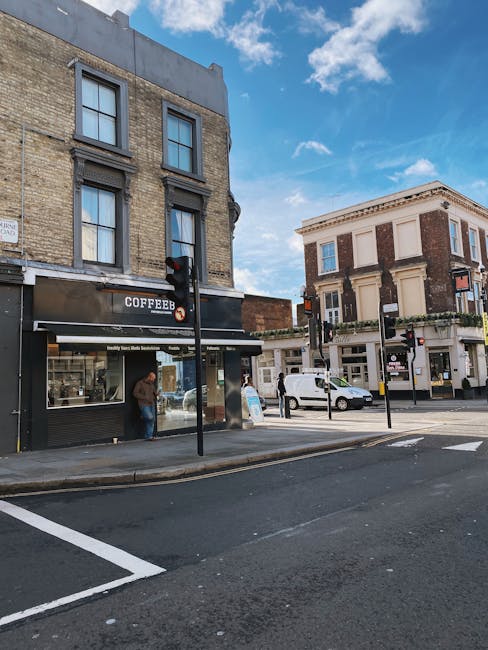 A street scene on Kentish Town Road showing a corner building with a cafe on the ground floor, featuring large glass windows and a black facade with a sign that reads 'Coffee'. Two people are visible inside, one near the window and the other standing outside on the sidewalk. A traffic light with red signal is positioned at the pedestrian crossing in front of the cafe. Parked cars are along the roadside, including a white van and a smaller vehicle. The buildings are historic-style, with brick facades and decorative architectural details, set against a partly cloudy blue sky. The scene is illuminated by daylight, highlighting the urban environment. This image relates to house removals and moving services in Kentish Town, where logistics and transport might involve navigating such streets when transporting furniture or belongings for a home relocation or office move, coordinated by Movers Kentish Town.