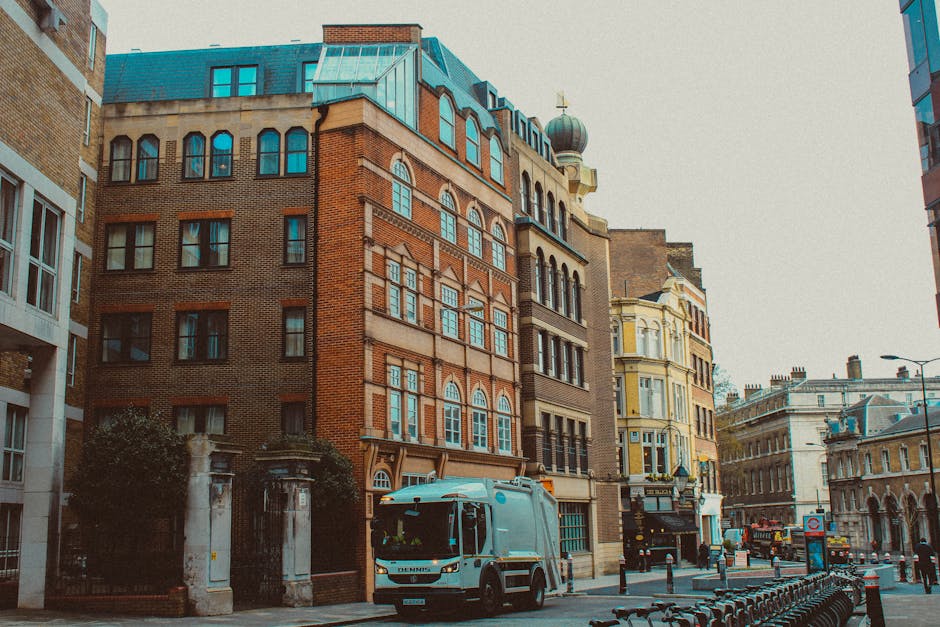 A street scene showing a modern multi-storey office building with large glass windows on the left and a historic curved sandstone building on the right near Kentish Town Road. In the center, there is a large tree with sparse green and brown leaves, indicating autumn. The street is paved with grey cobblestones, and several pedestrians are walking along the pavement. A white delivery van and a black car are parked near the buildings, with the van belonging to Movers Kentish Town, a professional removals company. Behind the vehicles, a loading area is visible with some caution cones and a few people standing nearby, possibly preparing for a house or office relocation. The overcast sky provides diffuse lighting, contributing to a neutral, daytime setting focused on urban moving logistics and furniture transport preparation in the vicinity of businesses and offices on Kentish Town Road.