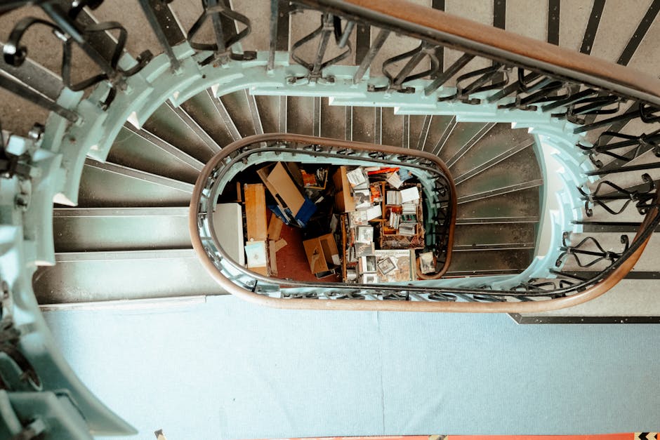 A top-down view of a spiral staircase inside a residential building, showing a wooden handrail, black metal balusters, and grey carpeted steps. The staircase curves downward, revealing a cluttered landing at the bottom with various cardboard boxes, books, and packing materials scattered across the floor. Some boxes are open, displaying contents such as books and small household items, while others are sealed or partially assembled. The stairs are well-lit with natural light coming from an unseen source above, illustrating the interior environment during a home relocation process. Occasionally, soft shadowed areas indicate the presence of moving equipment or personnel from Movers Kentish Town involved in the packing and furniture transport operation for a house or flat move with narrow staircases, typical in Kentish Town residences. The image emphasizes the careful loading process of packed boxes and furniture during an internal staircase move as part of a professional removals service.