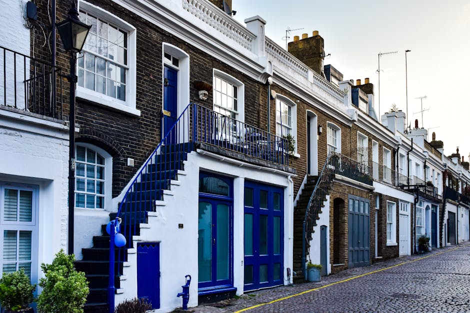 Exterior view of a row of terraced houses in Kentish Town, showcasing brick facades with white window frames and doors. The buildings feature small balconies with black and blue metal railings, accessible via external stairs with blue railings. The ground level includes large blue-framed windows and doorways, some covered with protective film or plastic wrapping, suggesting ongoing packing or moving activities. Potted plants and small shrubs are visible near the entrances. The cobbled sidewalk runs parallel to the houses, and a portion of a moving van or vehicle may be seen on the street. The scene illustrates a typical urban residential area that may be involved in home relocation or furniture transport, with external access points and narrow staircases suitable for a professional removals service like Movers Kentish Town conducting packing and loading operations.