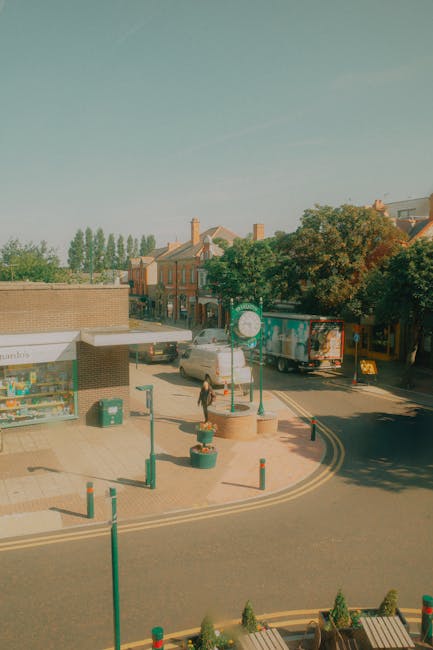 A street scene in a residential area with buildings and trees under clear blue sky. In the foreground, there is a small public square with a few green bollards, a recycling bin, and decorative flower pots. A woman carrying a cardboard box is walking on the pavement, approaching a white van parked at the curb, which is likely being used for home relocation or furniture transport. To the right, a larger truck with a colorful graphic design on its side is also parked, possibly involved in the moving process. The area is lit by natural daylight, with shadows cast by trees and vehicles, indicating it is daytime. This setting depicts an active loading or unpacking scene, consistent with moving or packing services provided by Movers Kentish Town, supporting their removals operations in the area around Fortess Road, Kentish Town NW5.