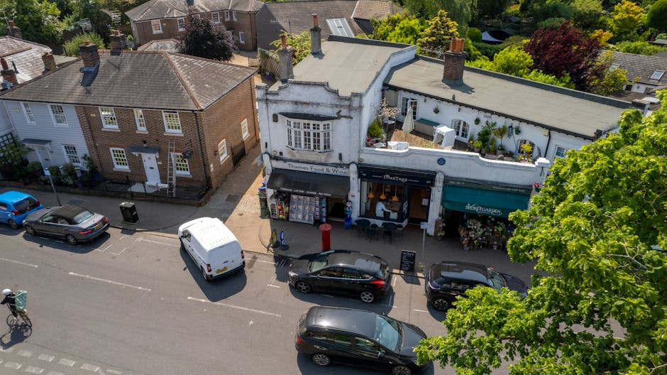 An aerial view of a quiet commercial street in Kentish Town, NW5, featuring a white two-story building with a rounded corner entrance that houses a small retail shop, currently displaying various merchandise outside on the sidewalk. Next to it, a single-story shop has a green awning and a display of potted plants. Several vehicles, including a white van, a black sedan, and a dark-colored hatchback, are parked along the street, with some space available for parking or loading. A bicycle is secured to a stand on the pavement, and the street is lined with trees providing shade and greenery. The area appears peaceful and suitable for local house removals or furniture transport, with the urban environment and surrounding residential buildings visible in the background. This scene exemplifies the typical setting for home relocation or moving services carried out by companies like Movers Kentish Town, ensuring smooth logistics during packing and loading processes.