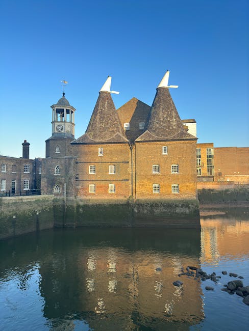 A historic brick building with two conical towers featuring white caps and small windows, situated along a riverbank under a clear blue sky. The building also has a clock tower with a weather vane on top, and large rectangular windows along its facade. In front, there is a calm water reflection of the structure, with a few rocks visible in the water near the bank. This setting illustrates the exterior of a heritage site, possibly featured in removals and relocation services by Movers Kentish Town, with the building positioned near a paved area suitable for loading or unloading household contents during a home relocation or furniture transport process.